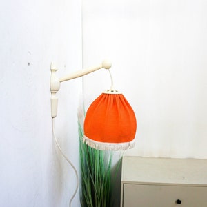 May include: A white wall-mounted lamp with a bright orange fabric shade. The shade has a white fringe trim.
