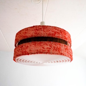 May include: A red and white pendant light fixture. The lampshade has a cylindrical shape with a textured red fabric exterior and a white, layered interior. A dark brown band accents the middle. The light hangs from a white cord.
