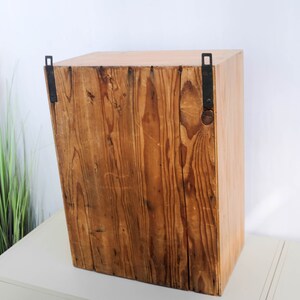 Op de afbeelding: Een houten kast met een rustieke bruine afwerking en zwarte metalen scharnieren. De kast heeft een eenvoudig ontwerp en is perfect voor het opbergen van kleine voorwerpen.