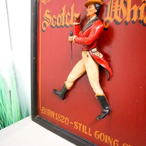 May include: A vintage wooden sign with a 3D figure of a man in a red jacket and white pants walking. The sign is red with gold lettering that reads "Scotch Whisky" and "Born 1820 - Still Going Strong".