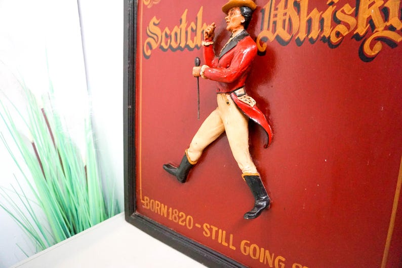 May include: A vintage wooden sign with a 3D figure of a man in a red jacket and white pants walking. The sign is red with gold lettering that reads "Scotch Whisky" and "Born 1820 - Still Going Strong".