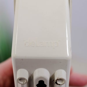 K&ouml;nnte beinhalten: Nahaufnahme einer wei&szlig;en Steckdose mit dem aufgedruckten Wort "delamp". Die Steckdose hat drei Stifte.