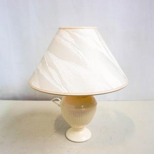 May include: A white table lamp with a cream-coloured shade featuring a subtle swirl pattern. The lamp base is a cream-coloured ceramic with a rounded shape.