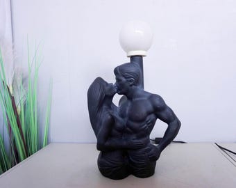 May include: A black sculpture of a couple kissing, with a white globe light above. The figures are embracing, with the man's muscular physique visible. The lamp is on a table.