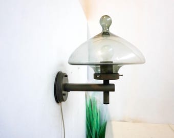 Può includere: Un'applique da parete in metallo grigio con un paralume in vetro trasparente. Il paralume ha la forma di un fungo e contiene una piccola lampadina. L'applique è montata su una parete bianca.