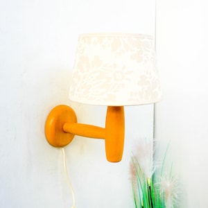 Może przedstawiać: Drewniana lampa ścienna z białym abażurem w kwiatowy wzór. Lampa ma prosty design z zakrzywionym ramieniem i okrągłą podstawą.