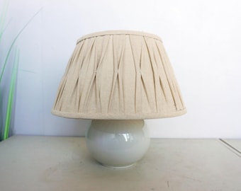 Puede incluir: Una pequeña lámpara de mesa color crema con una pantalla plisada. La pantalla se asienta sobre una base redonda y blanca. La lámpara está sobre una superficie clara, con plantas verdes en el fondo.