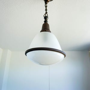Op de afbeelding: Een hanglamp met een klokvormige kap van matglas met een gladde witte afwerking. De armatuur is voorzien van een donkerbruine metalen kap en een bijpassende cirkelvormige band rond het midden, opgehangen aan een donkere metalen ketting.