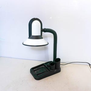 Puede incluir: Una lámpara de escritorio en blanco y negro con un cuello flexible verde y una base rectangular. La lámpara tiene una pantalla blanca con un borde negro y dos portalápices integrados. La lámpara está enchufada.