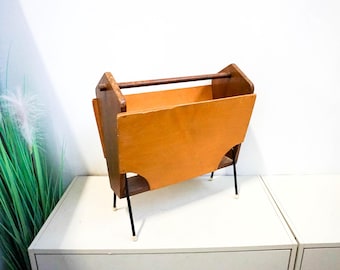 May include: A mid-century modern magazine rack made of wood with a light brown finish and black metal legs. The rack has a rectangular shape and is designed to hold magazines or other reading materials.