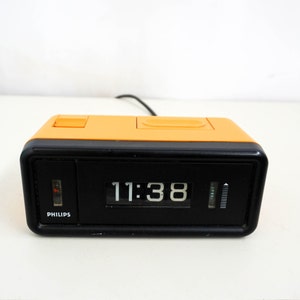 Può includere: Un orologio digitale nero e arancione con un display a lembo che mostra l'ora 11:38. L'orologio è etichettato "PHILIPS" sul davanti.