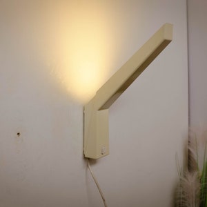 K&ouml;nnte beinhalten: Eine beige Wandleuchte mit rechteckigem Sockel und einem langen, abgewinkelten Arm. Das Licht ist eingeschaltet und strahlt ein warmes Licht ab.