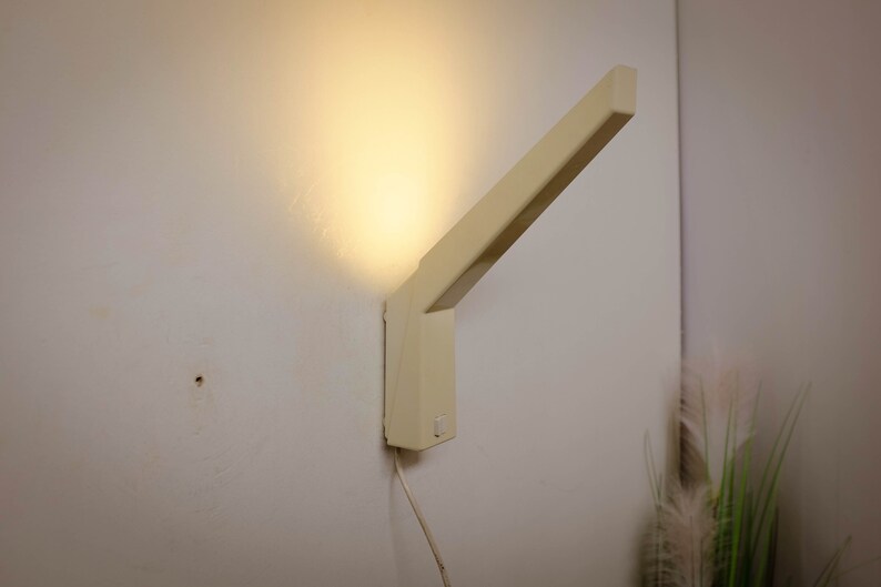 K&ouml;nnte beinhalten: Eine beige Wandleuchte mit rechteckigem Sockel und einem langen, abgewinkelten Arm. Das Licht ist eingeschaltet und strahlt ein warmes Licht ab.