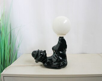May include: A black ceramic table lamp shaped like a person balancing a white globe on their head.