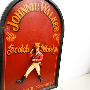 May include: A vintage wooden sign with a black frame and a red background. The sign features the text "Johnnie Walker Scotch Whisky" in gold lettering. A small, three-dimensional figure of a man in a red coat and top hat is standing on the sign. The text "Still Going Strong" is at the bottom of the sign.