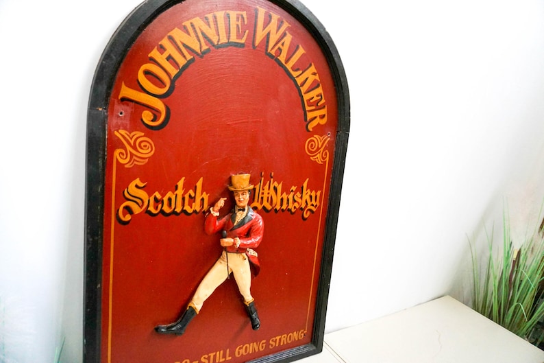 May include: A vintage wooden sign with a black frame and a red background. The sign features the text "Johnnie Walker Scotch Whisky" in gold lettering. A small, three-dimensional figure of a man in a red coat and top hat is standing on the sign. The text "Still Going Strong" is at the bottom of the sign.