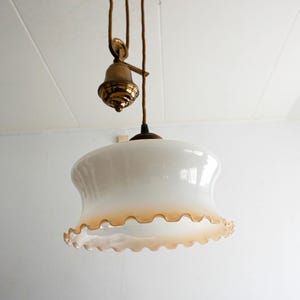 May include: Vintage white glass pendant light with a scalloped amber edge. The light fixture has a brass pulley system and is suspended from the ceiling. A classic lighting fixture.