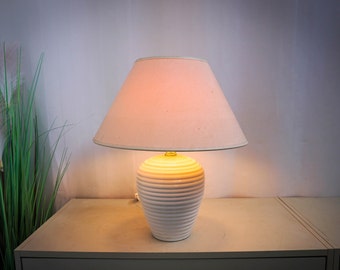 May include: A white ceramic table lamp with a cream lampshade. The lamp base features a ribbed design. The lamp is illuminated, producing a warm light. The lamp is placed on a light-coloured surface.