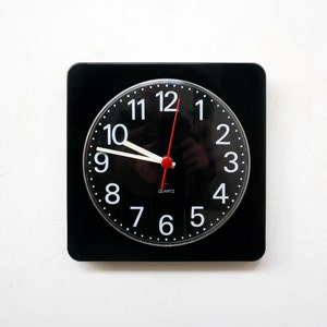 Op de afbeelding: Een zwarte vierkante klok met witte cijfers en wijzers. De wijzerplaat van de klok is zwart met witte cijfers en wijzers. De klok is ingesteld op 12:00 uur.