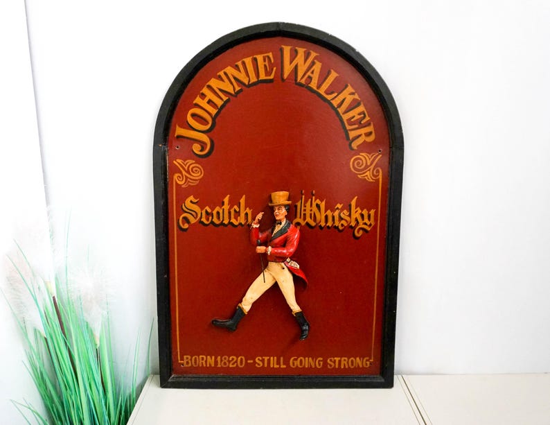 May include: A vintage wooden sign with a red background and gold lettering. The sign features a cartoon image of a man in a red coat and black top hat walking with a cane. The text on the sign reads "Johnnie Walker Scotch Whisky Born 1820 - Still Going Strong."