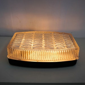 Peut inclure: Un luminaire de plafond rectangulaire avec un abat-jour en verre transparent orné d'un motif géométrique. La lumière est allumée et émet une lueur jaune chaude.