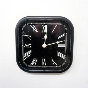 May include: A black square clock with Roman numeral markings and white hands. The clock face is black and the hands are white.