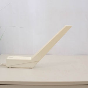 K&ouml;nnte beinhalten: Eine wei&szlig;e, eckige Tischlampe mit einem langen, d&uuml;nnen Arm. Die Lampe steht auf einer wei&szlig;en Oberfl&auml;che.