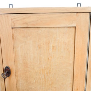 Op de afbeelding: Een licht houten kastje met een enkele deur en een sleutelgat. De kast heeft een eenvoudig ontwerp met een enkele plank binnenin.