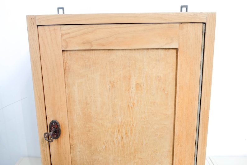 Op de afbeelding: Een licht houten kastje met een enkele deur en een sleutelgat. De kast heeft een eenvoudig ontwerp met een enkele plank binnenin.