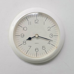 Op de afbeelding: Een witte analoge klok met zwarte wijzers en zwarte cijfers op een witte achtergrond. De wijzerplaat van de klok geeft "QUARTZ" aan.