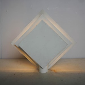 May include: White geometric table lamp with a layered diamond design. The lamp has a square base and a series of diamond-shaped layers, creating a modern, minimalist aesthetic. The lamp is illuminated from within, casting a warm glow.