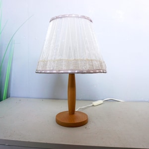 Op de afbeelding: Een tafellamp met een lichtgekleurde houten voet en een centrale standaard. De lamp heeft een geplooide, halfdoorzichtige witte stoffen kap met een decoratieve rand aan de onderkant. Een wit netsnoer met een schakelaar is bevestigd.