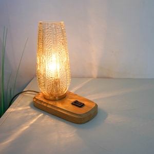 Könnte beinhalten: Eine Vintage-Tischlampe mit einem strukturierten Glasschirm und einem Holzsockel. Die Lampe hat eine Glühbirne im Inneren und ist eingeschaltet, wodurch ein warmes Licht erzeugt wird.