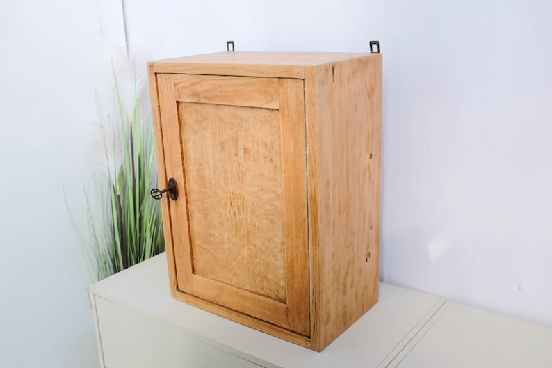 Op de afbeelding: Een klein houten kastje met een enkele deur en een sleutelgat. Het kastje is lichtbruin en heeft een eenvoudig ontwerp.