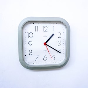 May include: A square-shaped, gray wall clock with white face and black numbers. The clock has a red second hand and black hour and minute hands. The clock face also has the word "quartz" printed on it.