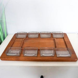 Op de afbeelding: Een houten serveerblad met acht kleine, heldere glazen schaaltjes. De lade is rechthoekig met een opstaande rand en een natuurlijke houten afwerking. De glazen schaaltjes hebben een vierkante vorm en een textuurpatroon.
