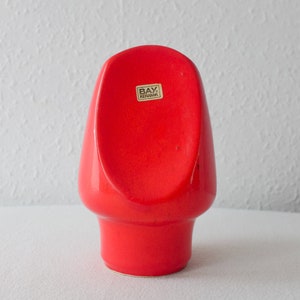 May include: A bright red ceramic vase with a unique, rounded shape. The vase has a label that reads "BAY Keramik".