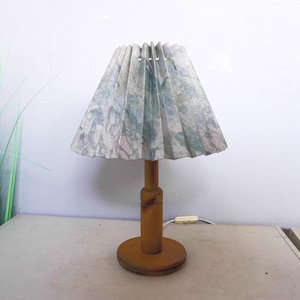Op de afbeelding: Een vintage houten tafellamp met een geplooide lampenkap. De lampenkap heeft een gemarmerd patroon in groen, blauw en beige. De lampvoet en -steel zijn gemaakt van lichtbruin hout. De lamp is aangesloten op een wit stopcontact.