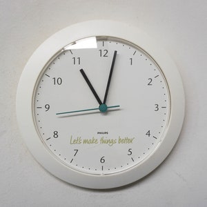 May include: White analogue clock with black hands and a teal second hand. The clock face has black numbers and the text "Let's make things better." in yellow.