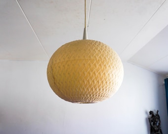 May include: A round, yellow pendant light fixture with a textured, woven pattern. The light hangs from a white ceiling with a thin, off-white cord. The fixture is a sphere shape, and the light is turned on.