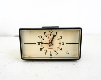 May include: A vintage rectangular clock with a black frame and a cream-colored face. The clock features black numerals, hands, and a red second hand. The clock face has the word "regotec" printed on it.