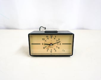 May include: A black alarm clock with a white face and black hands. The clock has a small light bulb icon on the face. The clock is set to 10:55.