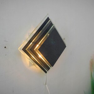 May include: A modern wall sconce with a geometric design. The sconce is made of layered glass and metal, and features a warm, white light.