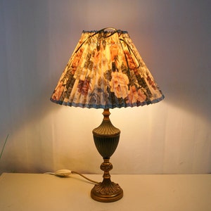 May include: A gold table lamp with a floral patterned lampshade. The lampshade is pleated and has a blue trim. The lamp is on a white surface.