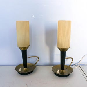 Op de afbeelding: Twee vintage tafellampen. Elke lamp heeft een cilindrische, geribbelde, crèmekleurige kap, een zwarte steel en een messing voet met een klein handvat. De lampen staan op een wit oppervlak, met een witte muur op de achtergrond.