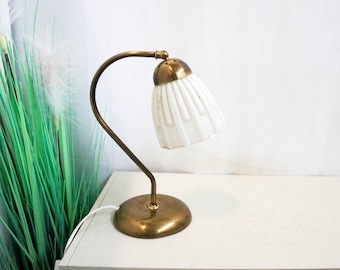 Peut inclure: Une lampe de bureau en laiton avec un abat-jour en verre plissé blanc. La lampe a un bras incurvé et une base ronde.