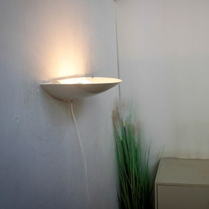 May include: A white wall-mounted light fixture with a curved, dish-shaped shade. The light is turned on and illuminates the wall. The fixture is wired with a white cord.
