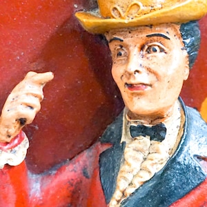 May include: A vintage figurine of a man wearing a yellow top hat, a black bow tie, and a red jacket. The figurine is pointing with his right hand.