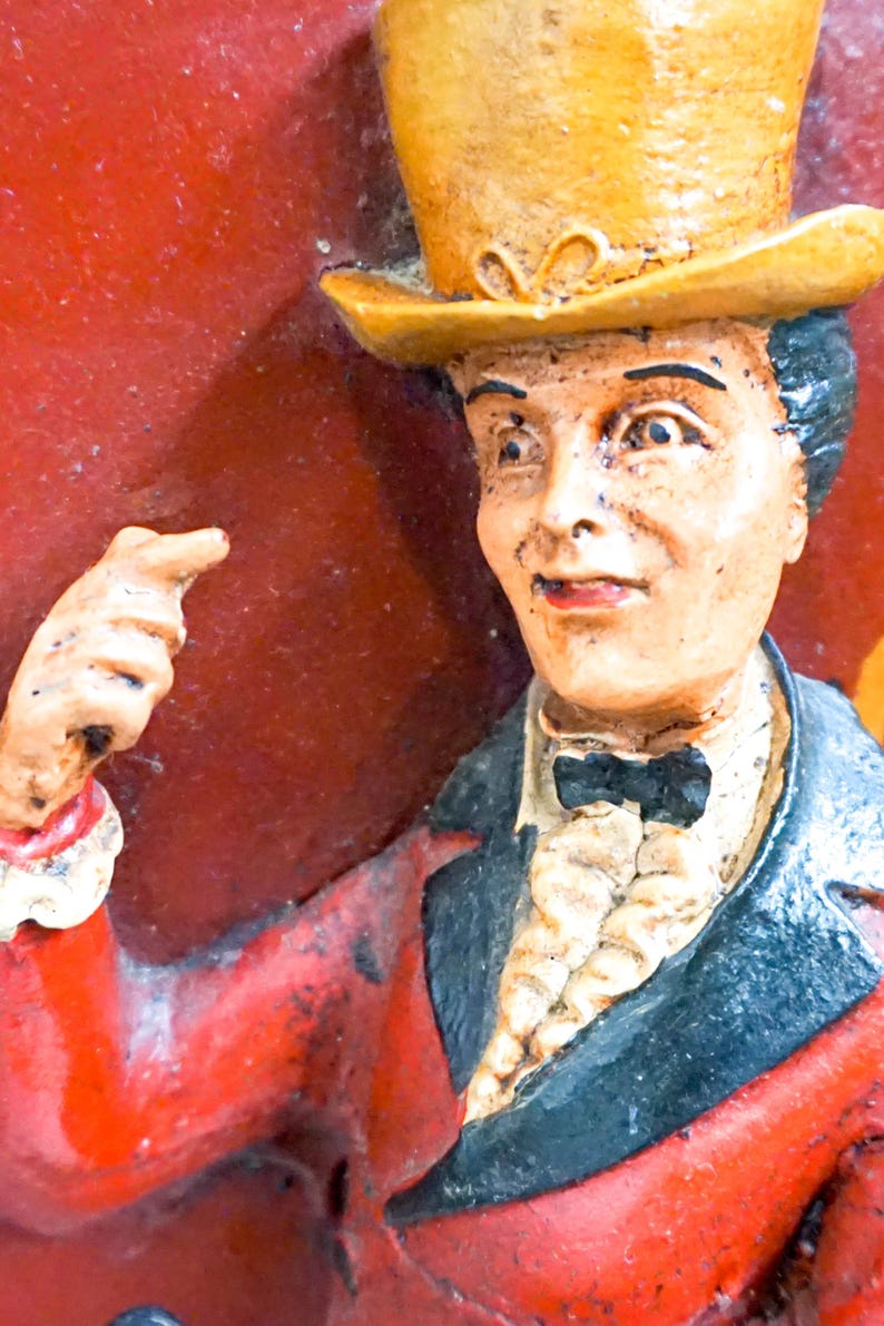 May include: A vintage figurine of a man wearing a yellow top hat, a black bow tie, and a red jacket. The figurine is pointing with his right hand.