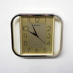 May include: A square, cream-coloured clock with black numbers and hands. The clock has a silver frame with a raised edge and is labelled "Academy Quartz".